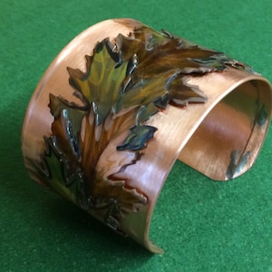 May include: Copper cuff bracelet with hand-painted leaves in shades of green, brown, and blue. The wide bracelet has a polished finish and a nature-inspired design. A unique piece of jewelry.