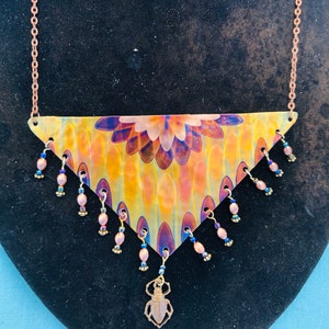 May include: A triangular copper necklace with a colorful iridescent design and a dangling beetle charm. The necklace features a beaded fringe with pink and blue beads.