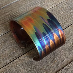 May include: A wide, curved cuff bracelet with a colorful, iridescent finish. The bracelet features a blend of orange, blue, green, and purple hues. The wood grain is visible on the inside of the bracelet.