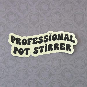 May include: A die-cut sticker with a cream-colored background and bold, black, wavy-style lettering that reads "PROFESSIONAL POT STIRRER". The sticker has a smooth, matte finish and features a distinct, irregular shape that follows the text.