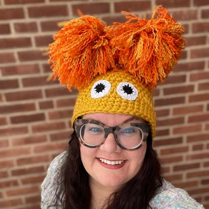 May include: A woman wearing a yellow crocheted hat with orange pom-poms and large white eyes. The hat has a black outline around the eyes.