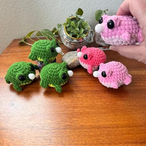 May include: Crocheted frog and axolotl plushies in shades of green and pink. The frog plushies have black eyes and white noses. The axolotl plushies are pink with black eyes and white noses. The plushies are displayed on a wooden surface.