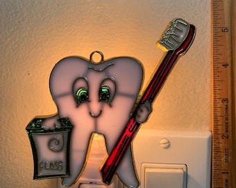 May include: A white tooth shaped night light with a red toothbrush and a green floss container. The tooth has green eyes and a smiling face. The night light is plugged into an electrical outlet.