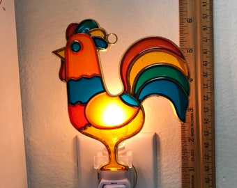 May include: A stained glass rooster nightlight with a yellow, orange, blue, green, and red color scheme. The rooster is mounted on a white plastic plug-in nightlight.