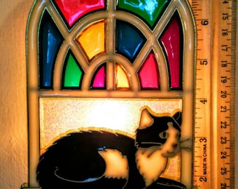 May include: A stained glass night light with a black and white cat silhouette inside. The stained glass features a rainbow of colors, including red, orange, yellow, green, blue, and purple. The night light is plugged into an electrical outlet.