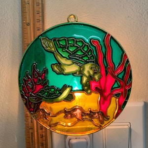 May include: A round, green, yellow, and red nightlight with a sea turtle swimming in front of coral. The nightlight is plugged into an electrical outlet.