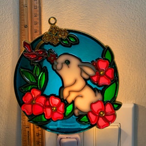 May include: A stained glass nightlight featuring a white bunny with red flowers and green leaves on a blue background. The nightlight is plugged into an electrical outlet.