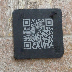 May include: A square, dark grey slate coaster featuring a light grey, laser-engraved QR code centred on its textured surface. The coaster has rough, uneven edges and a natural stone finish, providing a distinct contrast between the dark material and the light, geometric pattern of the code.