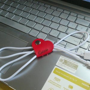 May include: A red heart-shaped leather cord tidy with the name "Lizzie" in cursive, secured with a silver popper. White USB charging cables are attached. The tidy is resting on a laptop.
