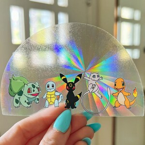Puede incluir: Una pegatina holográfica con cinco personajes de Pokémon: Bulbasaur, Squirtle, Umbreon, Eevee y Charmander. La pegatina tiene forma de semicírculo y es iridiscente.
