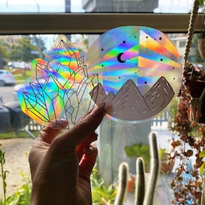 May include: Two iridescent holographic stickers. One sticker features a cluster of crystals. The other sticker features a mountain range with a crescent moon and stars.