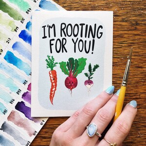 May include: A white card with the text "I'm rooting for you!" in black lettering. The card features a cartoon illustration of a carrot, a beet, and a turnip with faces.