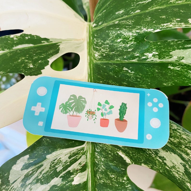 May include: A blue and white sticker of a Nintendo Switch console with a screen displaying five potted plants. The plants are a monstera, a hanging plant, a ficus, a succulent, and a cactus.
