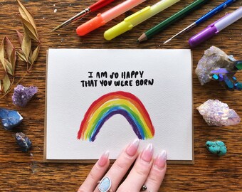 Könnte beinhalten: Eine Grußkarte mit einem Aquarell-Regenbogen und dem Text "I AM SO HAPPY THAT YOU WERE BORN." Die Karte liegt auf einer Holzoberfläche mit Kunstbedarf und Kristallen.