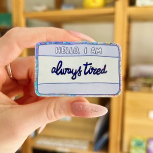 May include: A holographic sticker with a white background and blue text that reads "HELLO, I AM always tired".