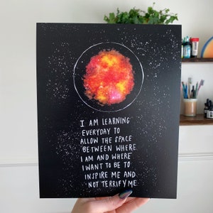 Könnte beinhalten: Schwarzer Kunstdruck mit einem farbenfrohen Himmelskörper in einem weißen Kreis, vor einem Sternenhintergrund. Der Druck enthält den Text: "I AM LEARNING EVERYDAY TO ALLOW THE SPACE BETWEEN WHERE I AM AND WHERE I WANT TO BE TO INSPIRE ME AND NOT TERRIFY ME."