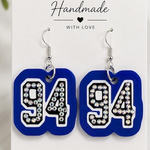 May include: A pair of blue dangle earrings featuring the number 94 in black with white borders, embellished with clear iridescent rhinestones. The earrings are displayed on a white card with the text Handmade with love and Thank you for supporting small business.