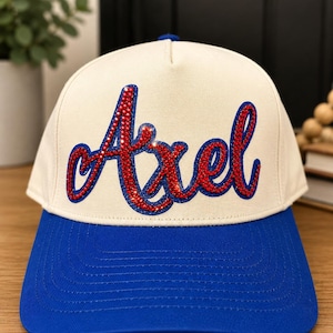 May include: A baseball cap with a cream-colored crown and a bright blue brim. The front of the crown features the name Axel in a cursive script, rendered in red rhinestones with a dark blue outline, creating a bold and textured appearance.