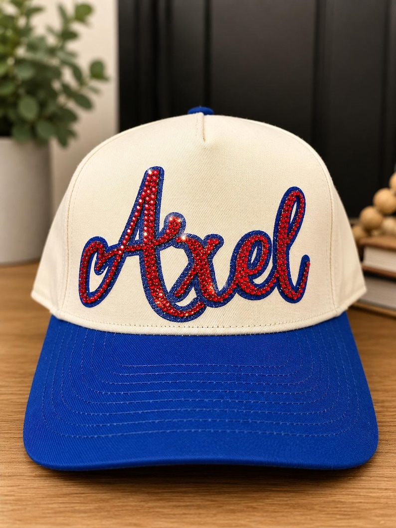 May include: A baseball cap with a cream-colored crown and a bright blue brim. The front of the crown features the name Axel in a cursive script, rendered in red rhinestones with a dark blue outline, creating a bold and textured appearance.