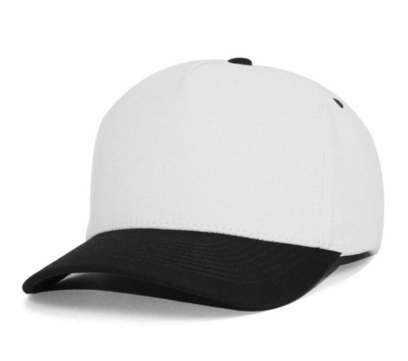 May include: A five-panel baseball cap featuring a solid white crown and a contrasting black curved brim. The cap includes a small black button at the top center and black eyelets for ventilation, providing a clean and structured appearance.