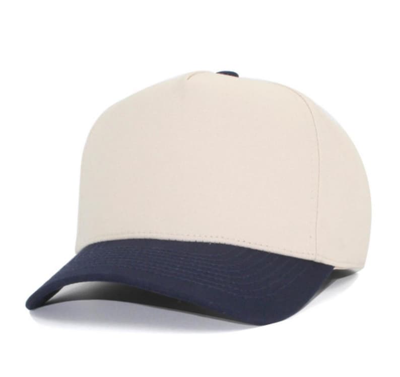 May include: A five-panel baseball cap featuring a solid cream-colored crown and a contrasting dark navy blue curved brim. The structured front panel is plain and unadorned, while the cap maintains a clean, simple design with visible stitching.