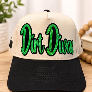 May include: A beige baseball cap with a black brim and a black button on top. The front features the text "Dirt Diva" in a cursive font, filled with bright green rhinestones and outlined in black, with black rhinestone accents on the side.