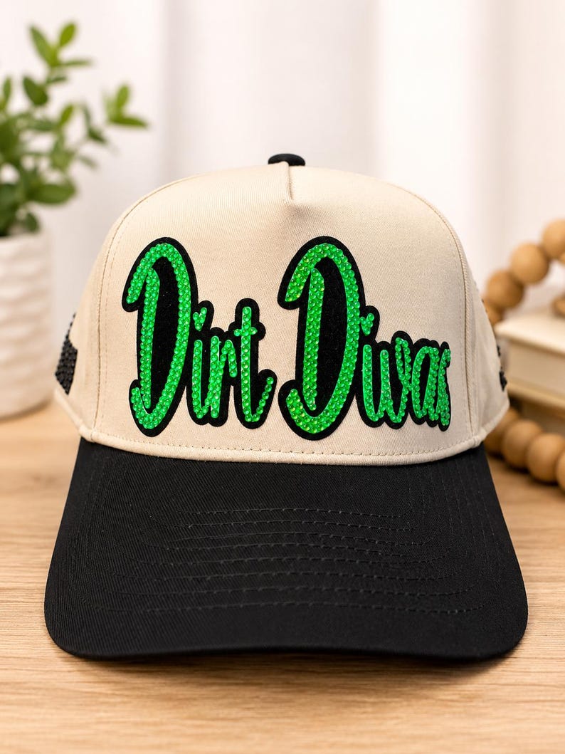 May include: A beige baseball cap with a black brim and a black button on top. The front features the text "Dirt Diva" in a cursive font, filled with bright green rhinestones and outlined in black, with black rhinestone accents on the side.