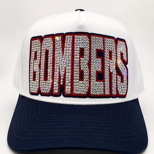 May include: A white baseball cap with a dark blue brim and a dark blue button on top. The front panel features the word BOMBERS in large, block letters filled with clear, reflective rhinestones and outlined in a thick dark blue and red border.