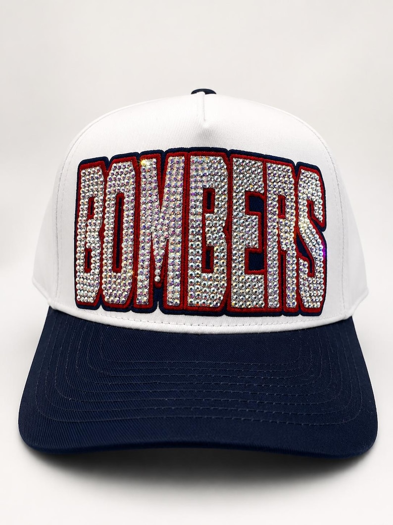 May include: A white baseball cap with a dark blue brim and a dark blue button on top. The front panel features the word BOMBERS in large, block letters filled with clear, reflective rhinestones and outlined in a thick dark blue and red border.
