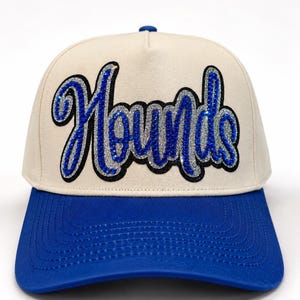 May include: A cream-colored baseball cap with a bright blue brim. The front panel features the word Hounds in a cursive script, rendered in blue rhinestones with a silver glitter outline and a black border, creating a textured, sparkling effect.
