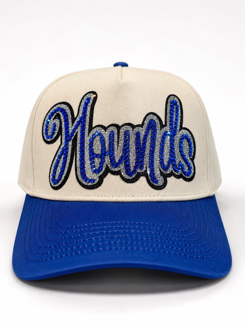 May include: A cream-colored baseball cap with a bright blue brim. The front panel features the word Hounds in a cursive script, rendered in blue rhinestones with a silver glitter outline and a black border, creating a textured, sparkling effect.