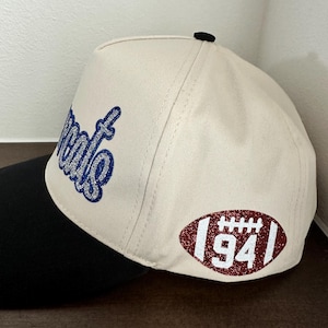 May include: A cream-colored baseball cap with a black brim and a black button on top. The side features a glittery blue script text and a glittery maroon football-shaped patch with white laces and the number 94 printed in the center.