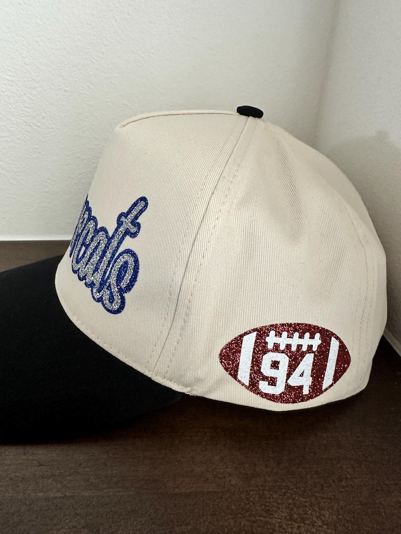 May include: A cream-colored baseball cap with a black brim and a black button on top. The side features a glittery blue script text and a glittery maroon football-shaped patch with white laces and the number 94 printed in the center.