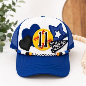 Könnte beinhalten: Eine blau-weiße Trucker-Cap mit einem gelben Softball-Aufnäher mit der Nummer 11, einem blauen Stern, zwei Herzen und einem Wimpel mit der Aufschrift GAME DAY. Eine Perlenschnur am Schirm zeigt den Text ROCK RIDGE mit kleinen Softball-Perlen.