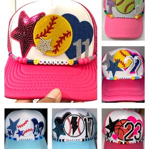 May include: Six different trucker hats with various sports themes. Each hat features a different sport, including baseball, softball, basketball, and lightning bolt. The hats are all white with a pink, blue, or white brim. Each hat has a different name on the front, including "Allyn", "Caden", and "Dello".