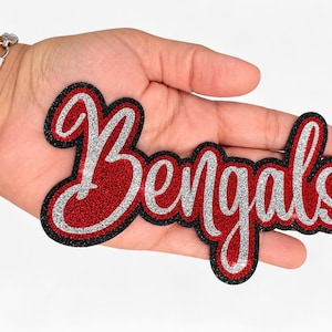 May include: A glittery patch featuring the word Bengals in a cursive script font. The lettering is silver with a red outline and a thick black border. The patch has a textured, sparkling finish throughout the entire design, creating a bold look.