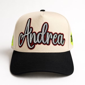 May include: A beige baseball cap with a black brim featuring the name Andrea in cursive script. The lettering is outlined in dark red and filled with clear, sparkling rhinestones. Bright neon yellow accents are visible on the side panels.