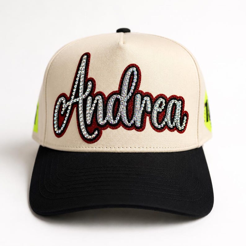 May include: A beige baseball cap with a black brim featuring the name Andrea in cursive script. The lettering is outlined in dark red and filled with clear, sparkling rhinestones. Bright neon yellow accents are visible on the side panels.
