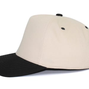 May include: A baseball cap featuring a cream-colored crown and a solid black brim. The cap includes a small black button at the top center and black eyelets on the side panels, providing a two-tone design with a structured front panel.