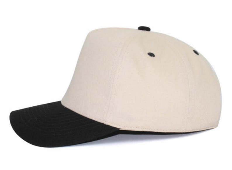 May include: A baseball cap featuring a cream-colored crown and a solid black brim. The cap includes a small black button at the top center and black eyelets on the side panels, providing a two-tone design with a structured front panel.