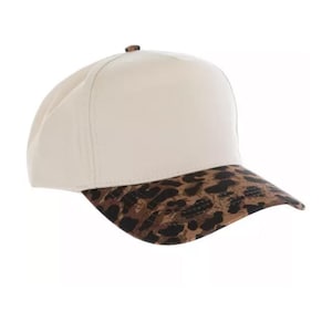 May include: A baseball cap featuring a solid cream-colored crown and a curved brim with a brown and black leopard print pattern. The cap has a small brown button at the top center and structured panels that meet at the crown of the head.