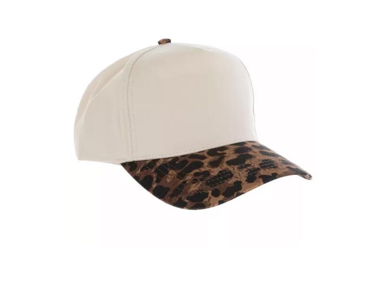 May include: A baseball cap featuring a solid cream-colored crown and a curved brim with a brown and black leopard print pattern. The cap has a small brown button at the top center and structured panels that meet at the crown of the head.