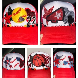May include: A collage of six red and white trucker hats with different sports-themed patches and embellishments. The hats feature a baseball, basketball, softball, and a megaphone. The hats also have beaded trim with the names "Olivia", "Woodlands Elite", and "Conrad" on them.