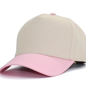 May include: A five-panel baseball cap featuring a solid beige crown and a contrasting light pink curved brim. The cap includes a small matching pink button at the top center and stitched detailing along the brim, providing a clean look.