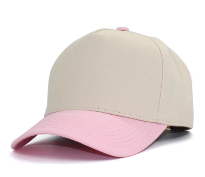 May include: A five-panel baseball cap featuring a solid beige crown and a contrasting light pink curved brim. The cap includes a small matching pink button at the top center and stitched detailing along the brim, providing a clean look.