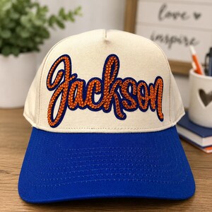 May include: A cream-colored baseball cap with a blue brim. The front panel features the name Jackson written in a cursive script, outlined in blue and filled with orange rhinestones that create a textured, sparkling appearance on the fabric.