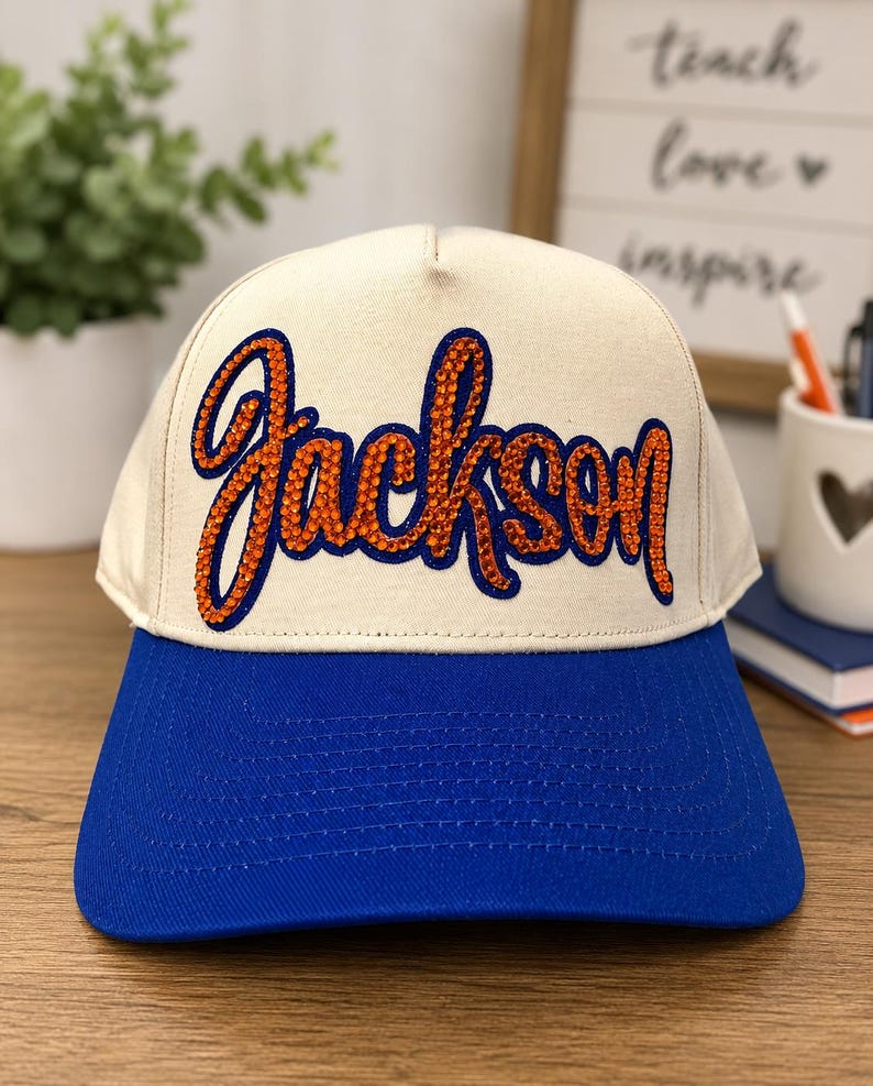 May include: A cream-colored baseball cap with a blue brim. The front panel features the name Jackson written in a cursive script, outlined in blue and filled with orange rhinestones that create a textured, sparkling appearance on the fabric.