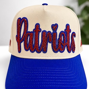 May include: A beige baseball cap with a bright blue brim and a blue button on top. The front features the word Patriots in red rhinestones with a blue rhinestone outline. The script lettering is bold and centered on the front panel of the cap.