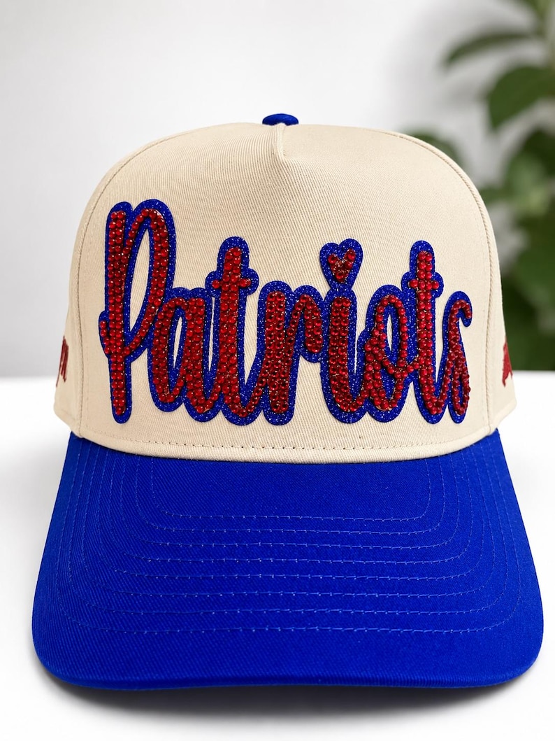 May include: A beige baseball cap with a bright blue brim and a blue button on top. The front features the word Patriots in red rhinestones with a blue rhinestone outline. The script lettering is bold and centered on the front panel of the cap.