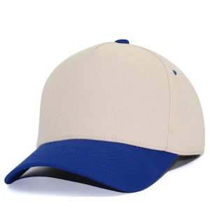 May include: A five-panel baseball cap featuring a solid cream-colored crown and a contrasting vibrant blue curved brim. The cap includes a small blue button at the top center and blue eyelets on the side panels, providing a clean and structured look.